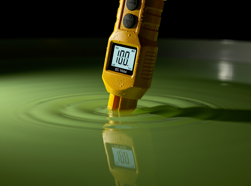 Yellow rugged truncheon-style EC meter probe being lowered into a hydroponic reservoir of green nutrient solution