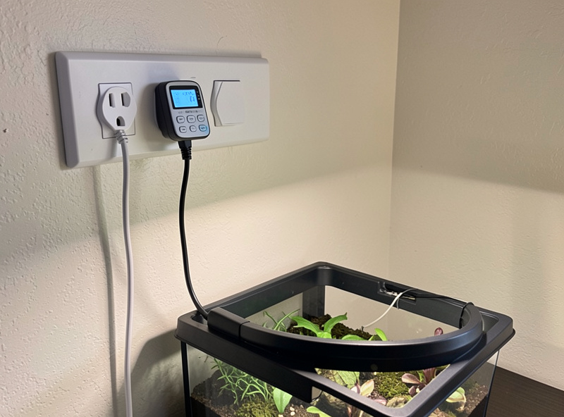 A programmable digital timer plugged into a power strip controlling a hydroponic grow light, with the reservoir visible below